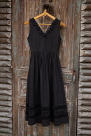 A sleeveless black midi dress with lace trim at the V-neckline and hem, hanging on a wooden hanger against a rustic weathered wooden door.