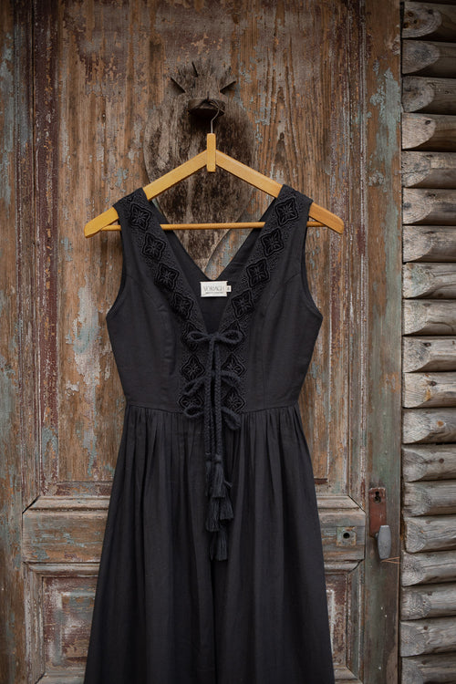 A sleeveless black midi dress with a lace-trimmed V-neckline and front tie detail, hanging on a wooden hanger against a rustic weathered wooden door.