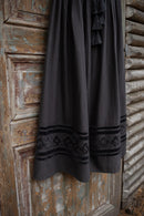 A dark charcoal gray linen dress with black embroidered geometric patterns and tassel ties, hanging against a rustic, weathered wooden door.