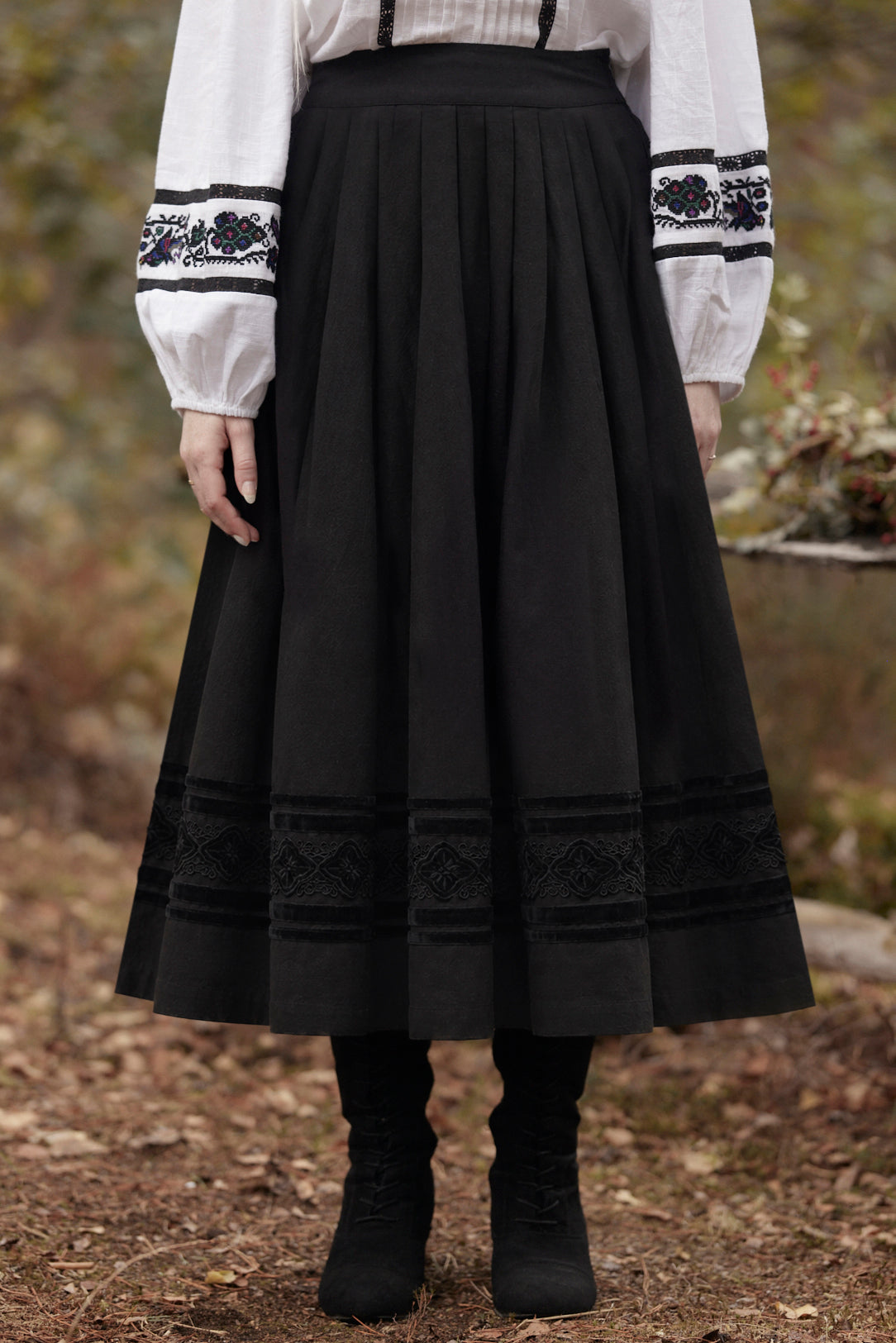 A person wearing a long black pleated skirt with embroidered trim at the hem and waistband, paired with a white blouse and black boots, standing outdoors on a forest floor.