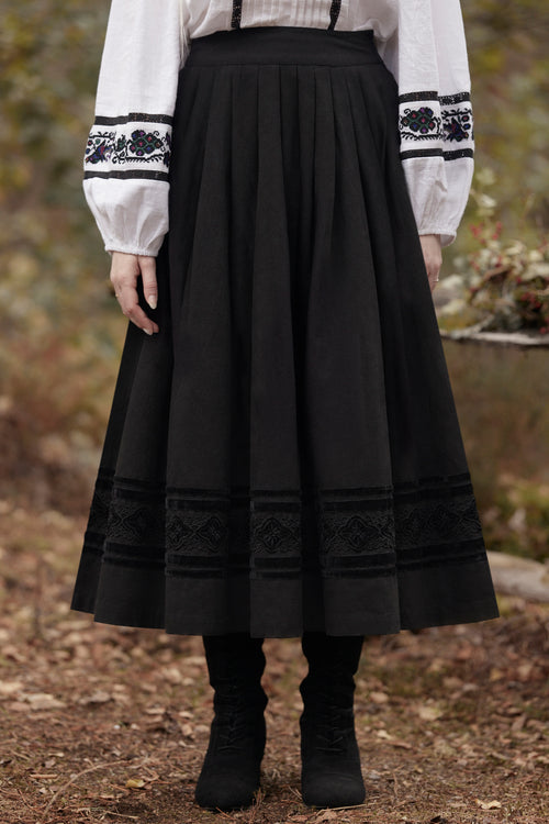 A person wearing a long black pleated skirt with embroidered trim at the hem and waistband, paired with a white blouse and black boots, standing outdoors on a forest floor.
