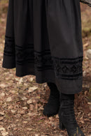 A close-up of a person wearing a long black skirt with embroidered trim and black lace-up ankle boots, standing outdoors on a leaf-covered ground.