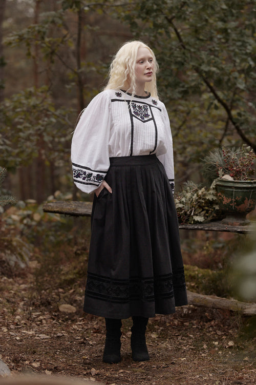 A woman with platinum blonde hair stands outdoors in a wooded setting wearing a white long-sleeve blouse with black embroidered details and a long black skirt.