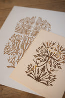 Two botanical art prints with intricate gold linocut-style illustrations on cream and white paper, arranged diagonally on a light wood surface.