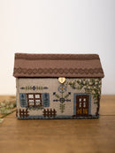 A small house-shaped needlepoint canvas with a brown roof, beige wall, blue shutters, a brown door, and decorative floral details, displayed on a wooden surface against a light background.