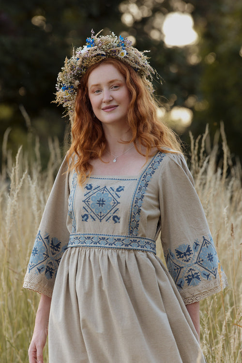 Woman in a beige dress with blue embroidery standing in a field with a floral crown.