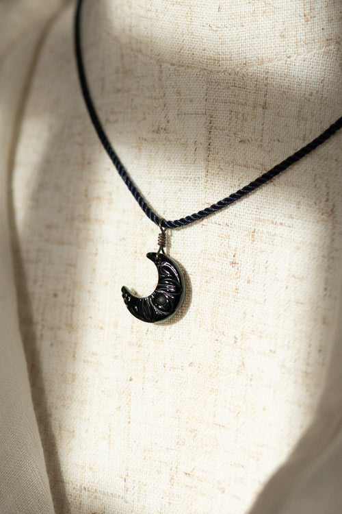 A black pendant necklace featuring a crescent moon charm with carved details, displayed on a textured beige fabric mannequin bust.