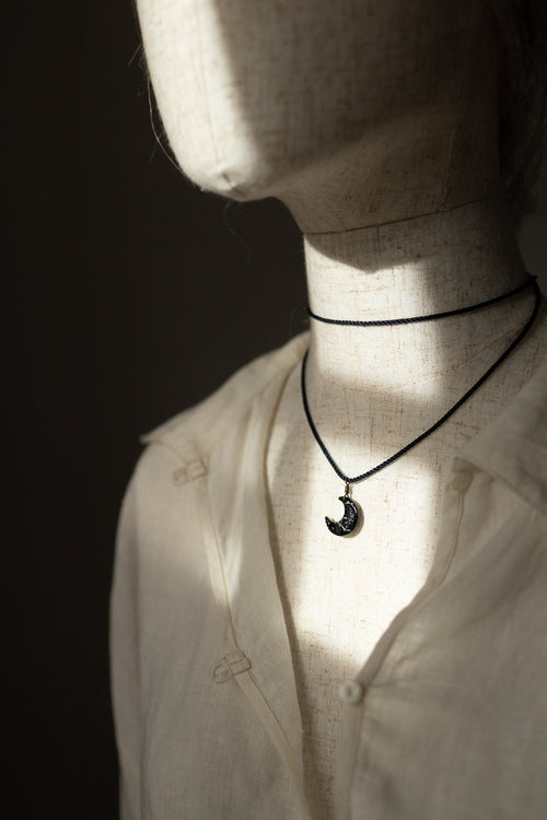 A black cord necklace with a crescent moon pendant displayed on a mannequin torso wearing a white shirt, lit by dramatic side lighting against a dark background.