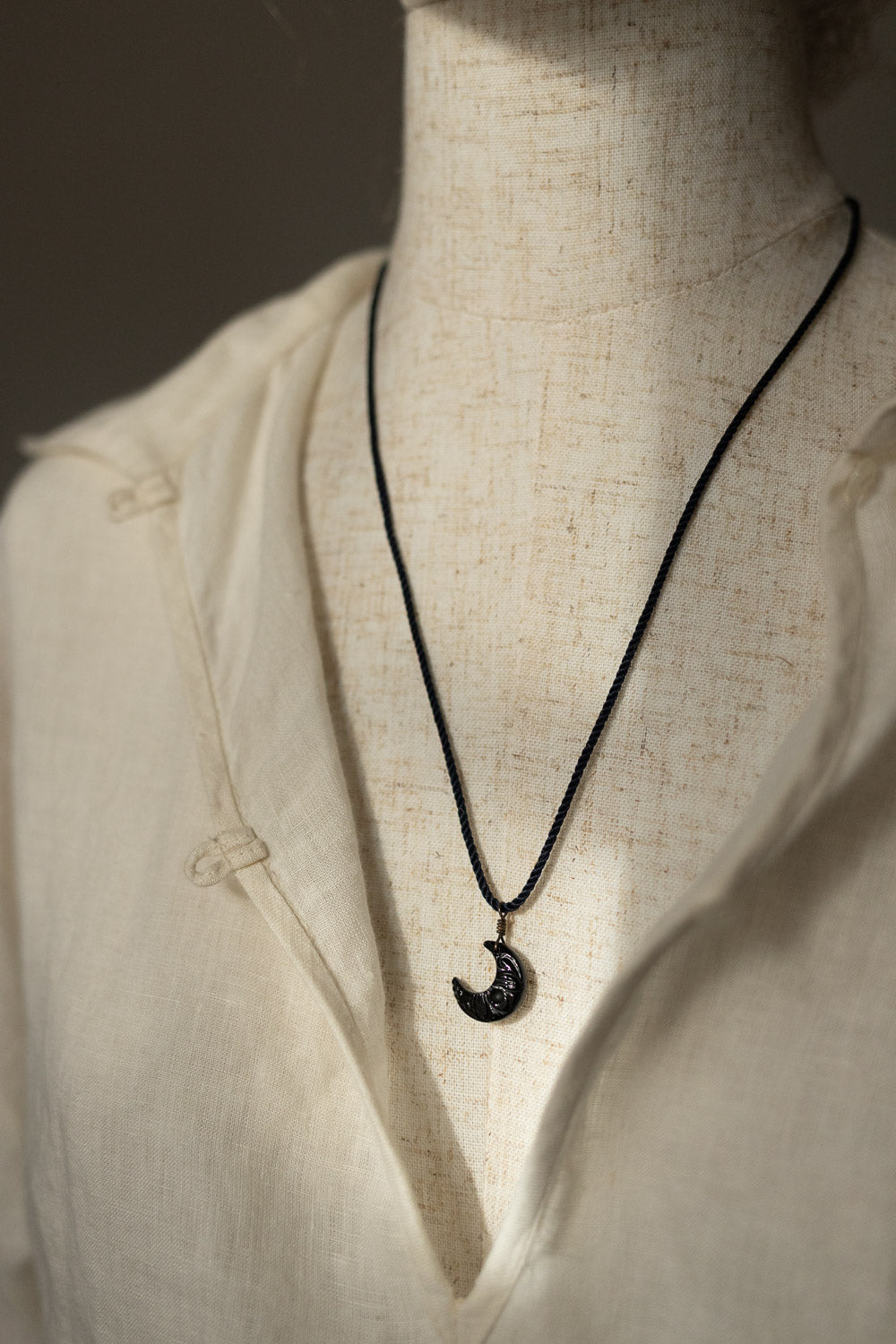 A black cord necklace with a crescent moon pendant featuring a textured black-and-white pattern, displayed on a cream linen shirt mannequin.