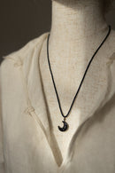 A black cord necklace with a crescent moon pendant featuring a textured black-and-white pattern, displayed on a cream linen shirt mannequin.