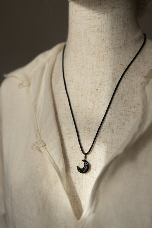 A black cord necklace with a crescent moon pendant featuring a textured black-and-white pattern, displayed on a cream linen shirt mannequin.