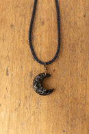 A black carved crescent moon pendant necklace on a black braided cord, displayed on a warm wooden surface.