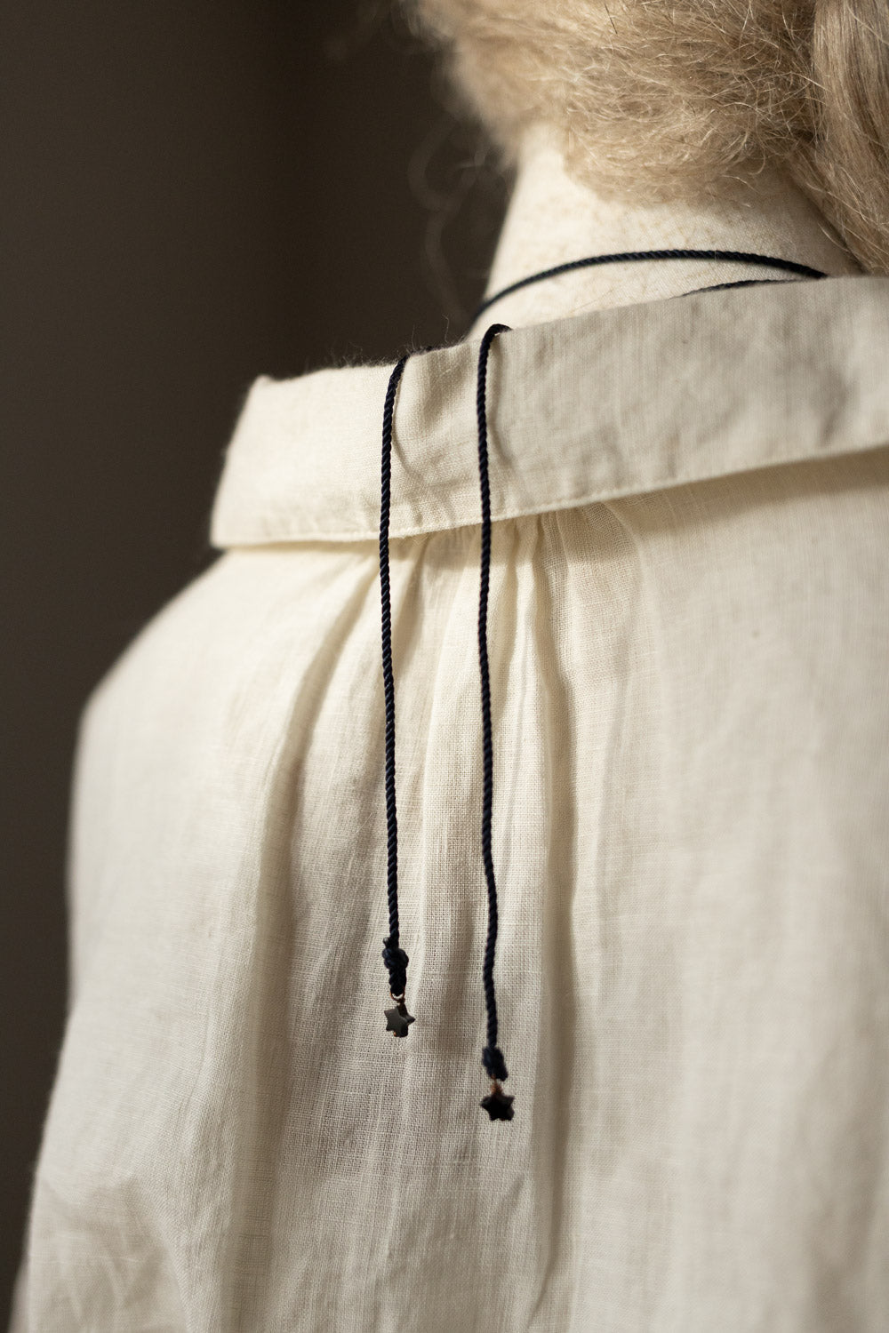 Close-up of the back of a cream-colored linen shirt with a collar, featuring two black drawstrings with small black cross pendants hanging down the center.