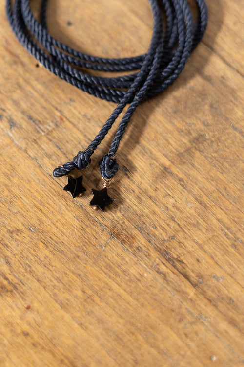 A close-up of a dark charcoal gray braided cord necklace with two small black star charms, resting on a warm brown wooden surface.