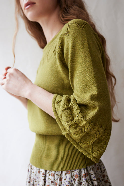 Woman wearing a green sweater with intricate sleeve detailing against a neutral background