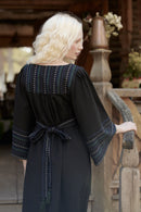A woman with long blonde hair is shown from the back wearing a black dress with blue and white embroidered details, standing next to a rustic wooden railing.