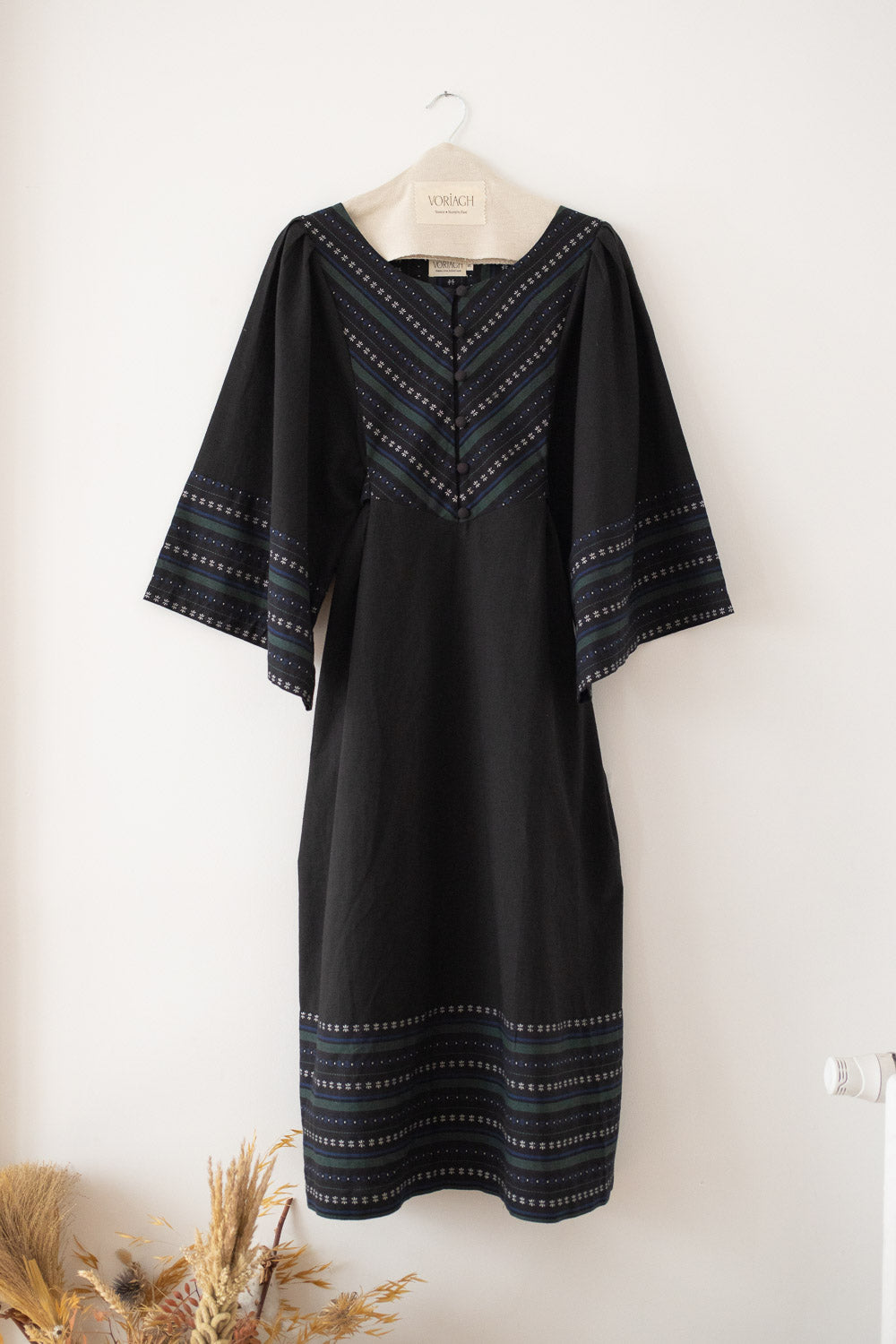 A black kaftan dress with blue and white geometric embroidered patterns on the neckline, sleeve cuffs, and hem, hanging on a wooden hanger against a white wall.