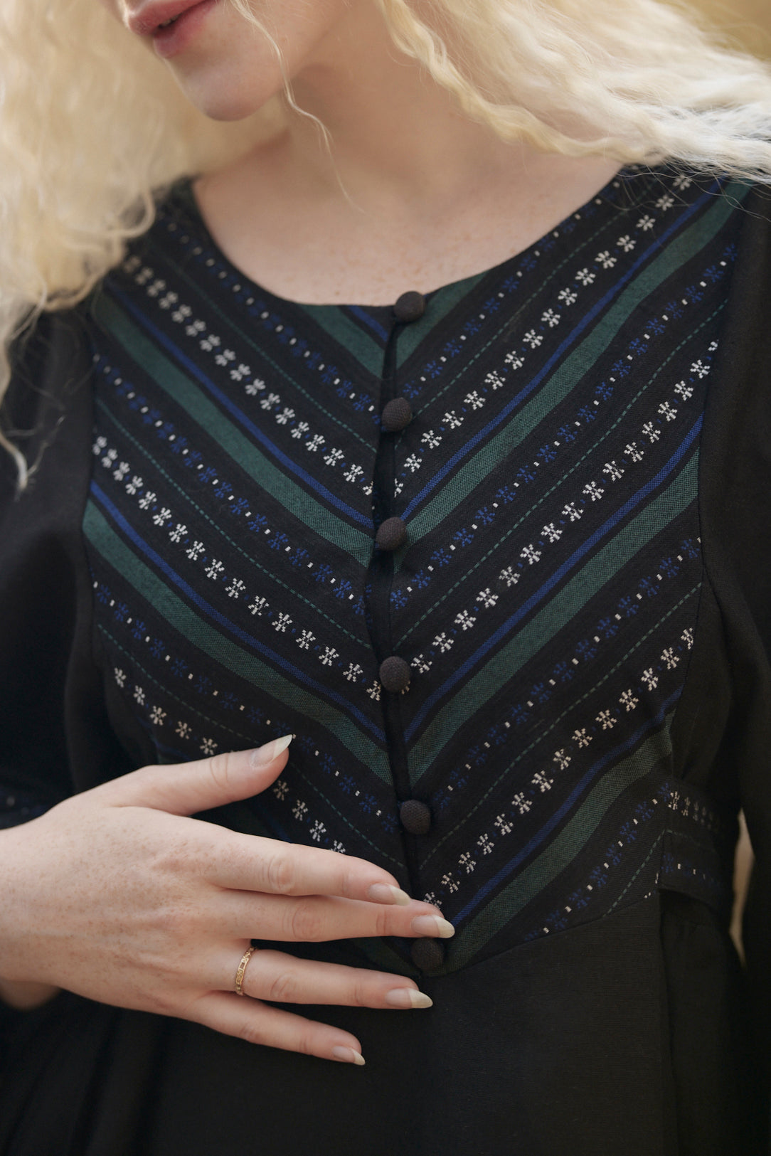 Close-up of a woman wearing a black dress with a decorative chest panel featuring diagonal stripes in dark green, navy, and white with star-like patterns and a button closure.