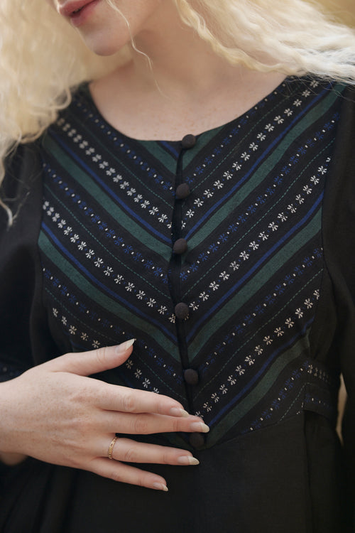 Close-up of a woman wearing a black dress with a decorative chest panel featuring diagonal stripes in dark green, navy, and white with star-like patterns and a button closure.