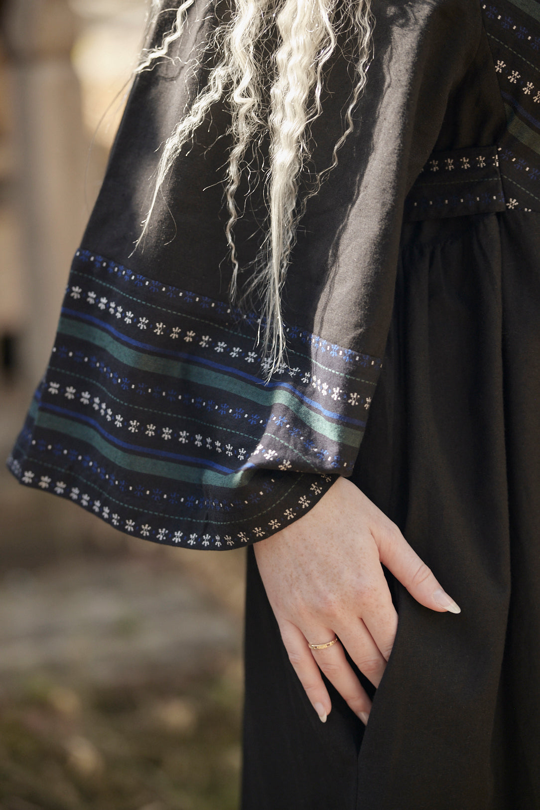 Close-up of a person wearing a black dress with wide sleeves featuring blue, green, and white striped patterns and star motifs near the cuffs. The person has long, wavy platinum blonde hair and is shown from the chest down, with one hand resting on the skirt.