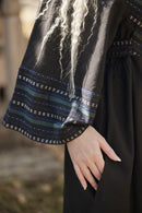 Close-up of a person wearing a black dress with wide sleeves featuring blue, green, and white striped patterns and star motifs near the cuffs. The person has long, wavy platinum blonde hair and is shown from the chest down, with one hand resting on the skirt.