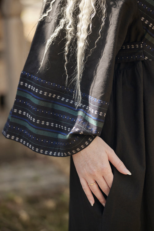 Close-up of a person wearing a black dress with wide sleeves featuring blue, green, and white striped patterns and star motifs near the cuffs. The person has long, wavy platinum blonde hair and is shown from the chest down, with one hand resting on the skirt.