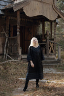 A woman with long platinum blonde hair stands in front of a rustic wooden cabin wearing a black long-sleeve dress with blue geometric patterns on the neckline and hem, paired with black boots.