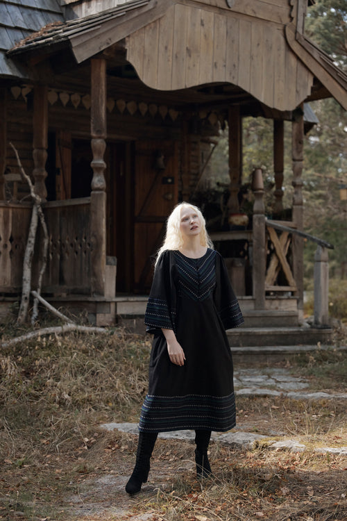 A woman with long platinum blonde hair stands in front of a rustic wooden cabin wearing a black long-sleeve dress with blue geometric patterns on the neckline and hem, paired with black boots.