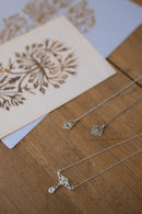 Two silver necklaces with geometric cutout pendants are displayed on a wooden surface next to a decorative card.