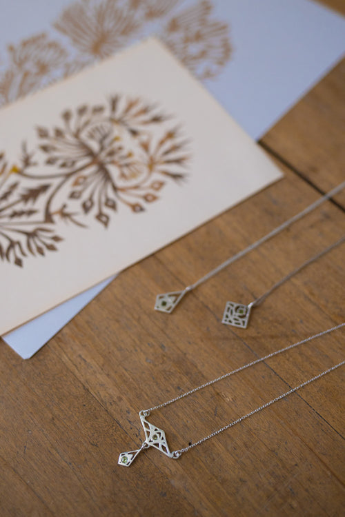 Two silver necklaces with geometric cutout pendants are displayed on a wooden surface next to a decorative card.
