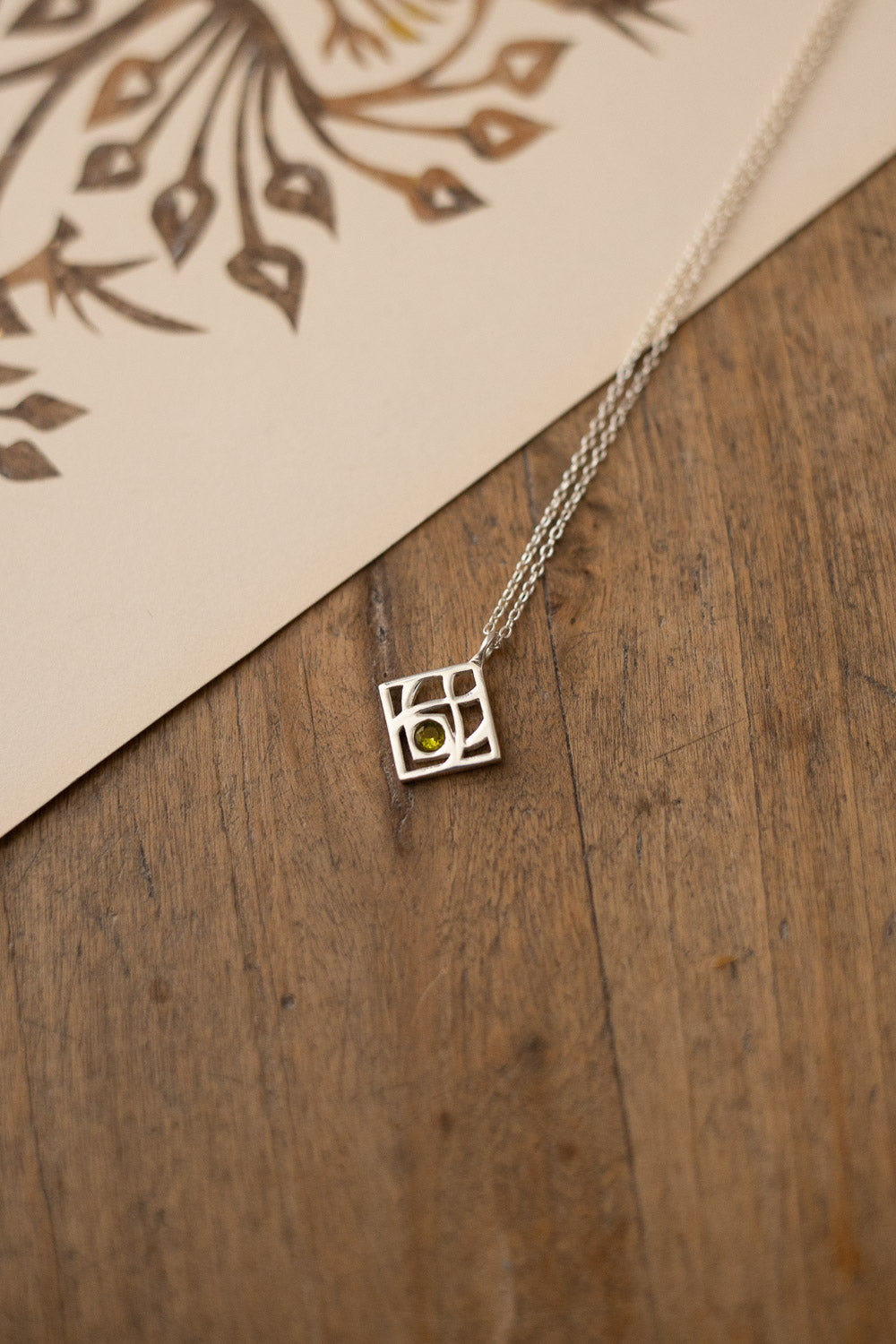 A silver geometric square pendant necklace with a small green stone at its center, resting on a rustic wooden surface next to a cream-colored card with a brown leaf pattern.