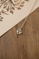 A silver geometric square pendant necklace with a small green stone at its center, resting on a rustic wooden surface next to a cream-colored card with a brown leaf pattern.
