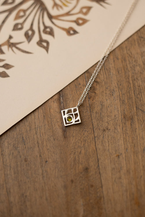 A silver geometric square pendant necklace with a small green stone at its center, resting on a rustic wooden surface next to a cream-colored card with a brown leaf pattern.