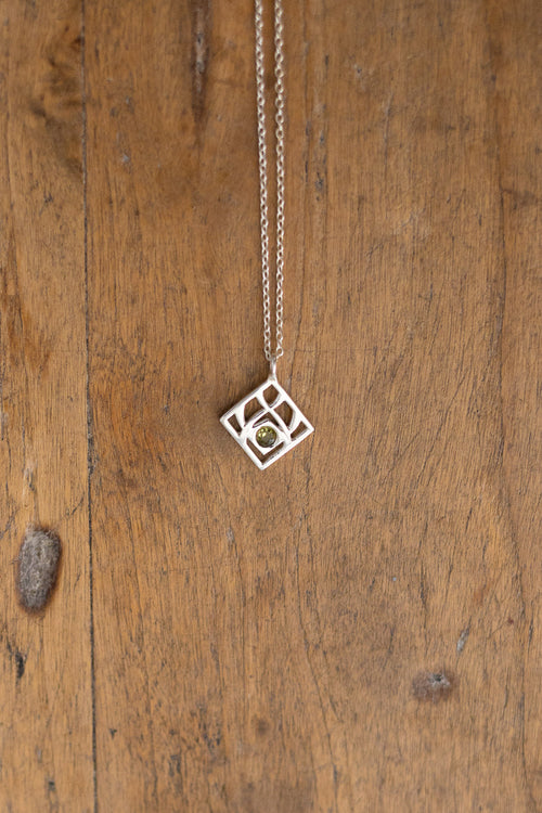A delicate silver geometric pendant necklace with a small central gold circle, hanging against a rustic wooden plank background.