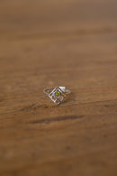 A silver geometric ring with a small green gemstone in the center, resting on a rustic wooden surface.