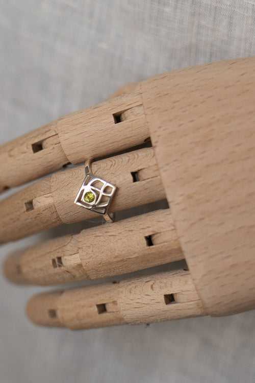 A silver ring with a geometric frame and a small green gemstone, displayed on a wooden mannequin hand against a neutral gray background.