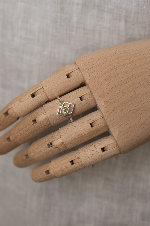 A silver ring with a small yellow-green gemstone set in a geometric square frame, displayed on a wooden mannequin hand against a textured gray fabric background.