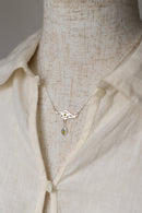 A delicate silver necklace with a geometric pendant and a small green teardrop charm, displayed on a textured cream linen mannequin torso wearing a matching cream linen shirt.