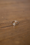 A delicate silver ring with a small central yellow gemstone and subtle side details, resting on a warm, blurred wooden surface.