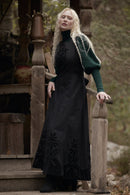 A woman with long platinum blonde hair stands on rustic wooden steps wearing a long black dress with floral embroidery and a dark green long-sleeve top underneath.