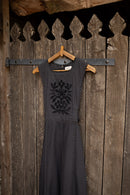 A dark charcoal gray sleeveless dress with a tonal floral embroidery on the chest, hanging on a wooden hanger against a rustic dark wood plank wall.