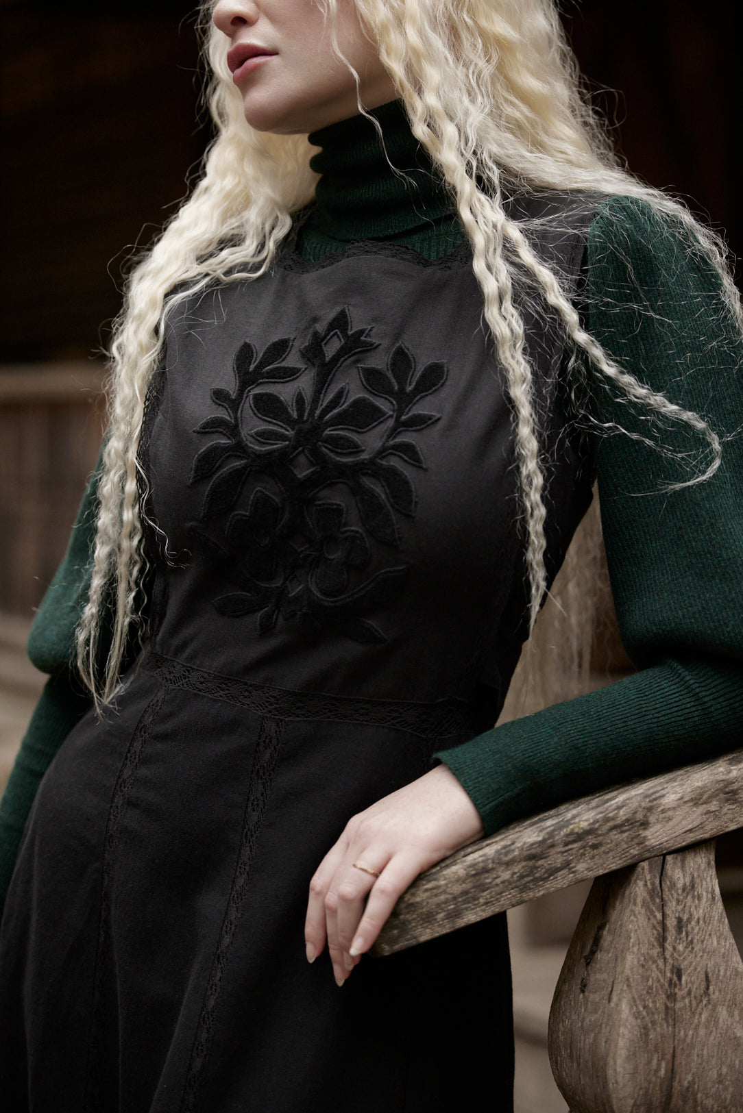 A close-up lifestyle shot of a model with long blonde braids wearing a black embroidered turtleneck dress over a dark green knit turtleneck sweater, leaning on a rustic wooden railing.