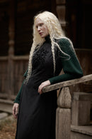 A woman with long platinum blonde hair wearing a black long-sleeve dress with a dark green knit sweater layered over the shoulders, leaning on a rustic wooden railing in front of a dark wooden building.