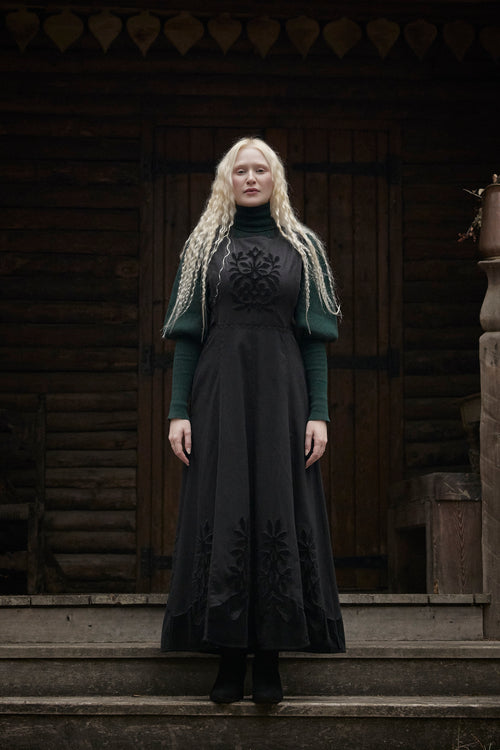 A woman with long blonde braided hair stands on wooden steps in front of a rustic log cabin doorway, wearing a long black dress with dark green sleeves and black lace embroidery.