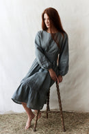 Woman in a gray dress sitting on a wooden stool against a white wall.