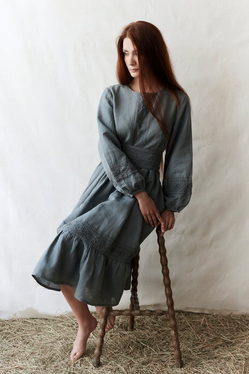 Woman in a gray dress sitting on a wooden stool against a white wall.