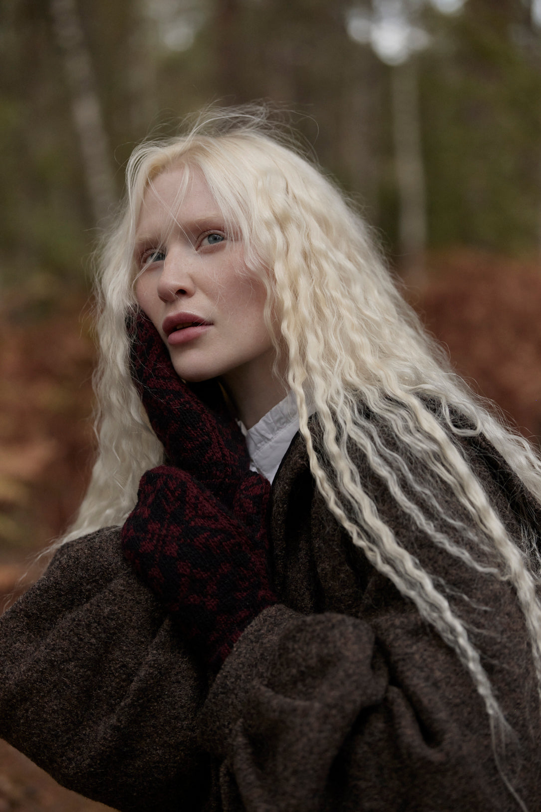 A woman with long, wavy platinum blonde hair wearing a dark brown textured coat, a dark red knit scarf, and a white shirt, photographed outdoors in a blurred forest setting.