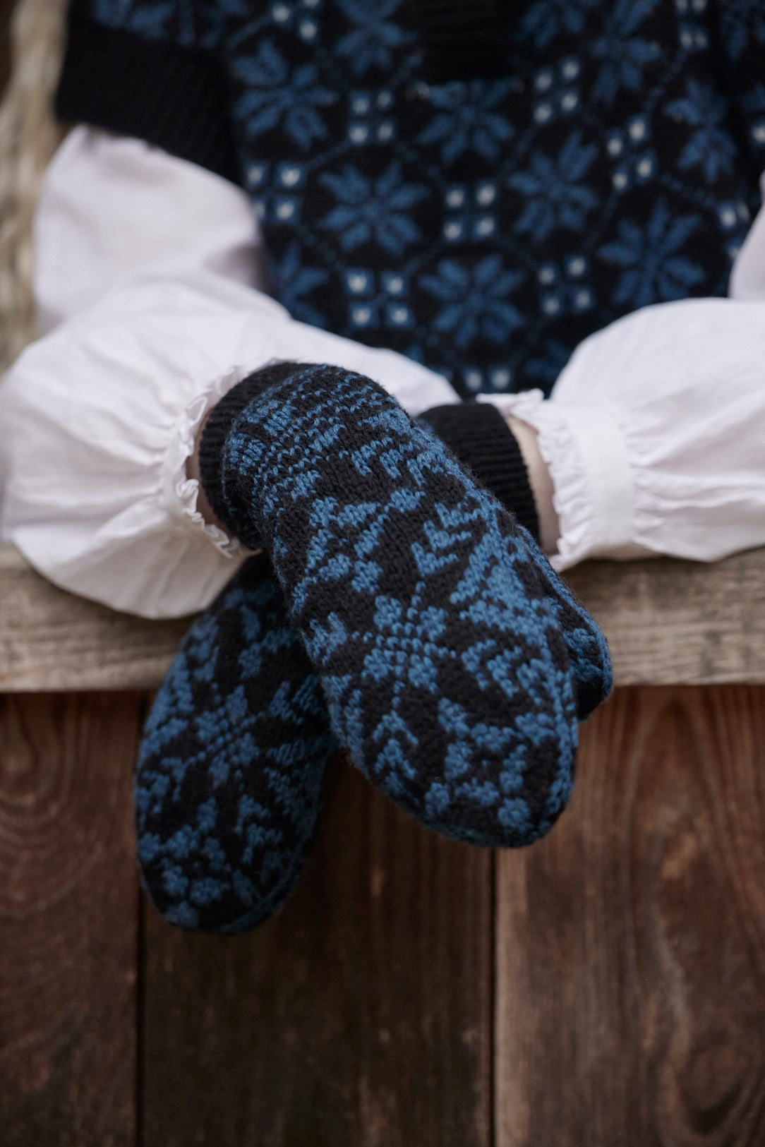A close-up of a person sitting on a wooden bench wearing dark navy and blue patterned knit mittens over white puffed sleeves and a matching dark sweater.