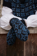 A close-up of a person sitting on a wooden bench wearing dark navy and blue patterned knit mittens over white puffed sleeves and a matching dark sweater.