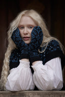 A young woman with long blonde hair wearing a dark navy blue and black patterned knit sweater vest over a white ruffled blouse, holding her hands to her cheeks with matching knit mittens.